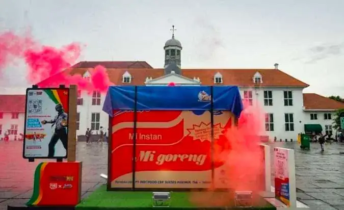 The Indomie Mi Goreng airdrop crate on green turf in Kota Tua square, Jakarta, with red smoke drifting past the old town hall
