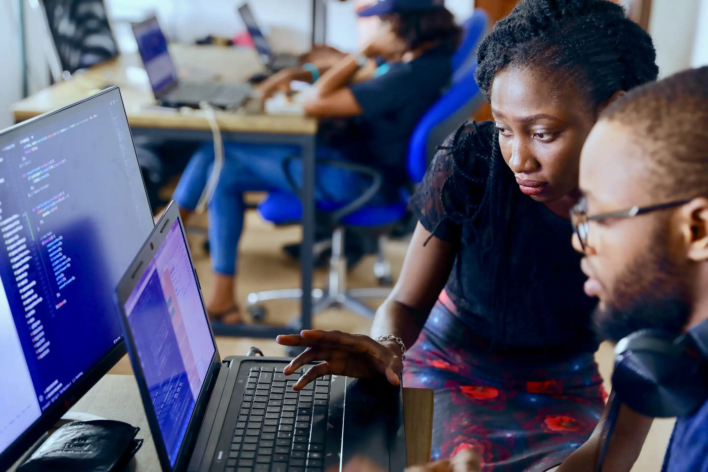 Two software developers look closely at a laptop screen beside a monitor full of code, with colleagues working in the background