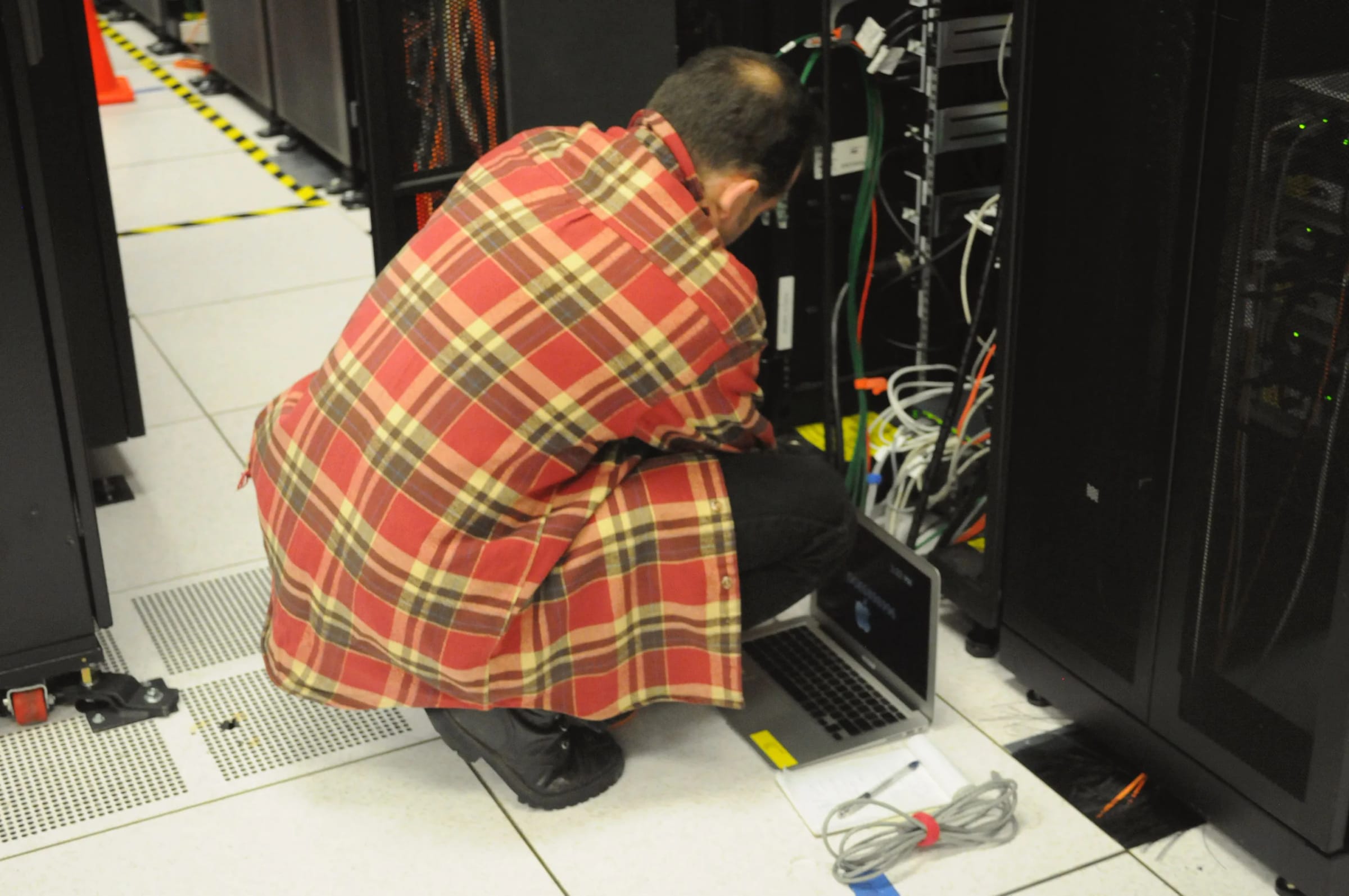 A technician crouches with a laptop on the floor at an open server rack, between rows of black cabinets in a data center