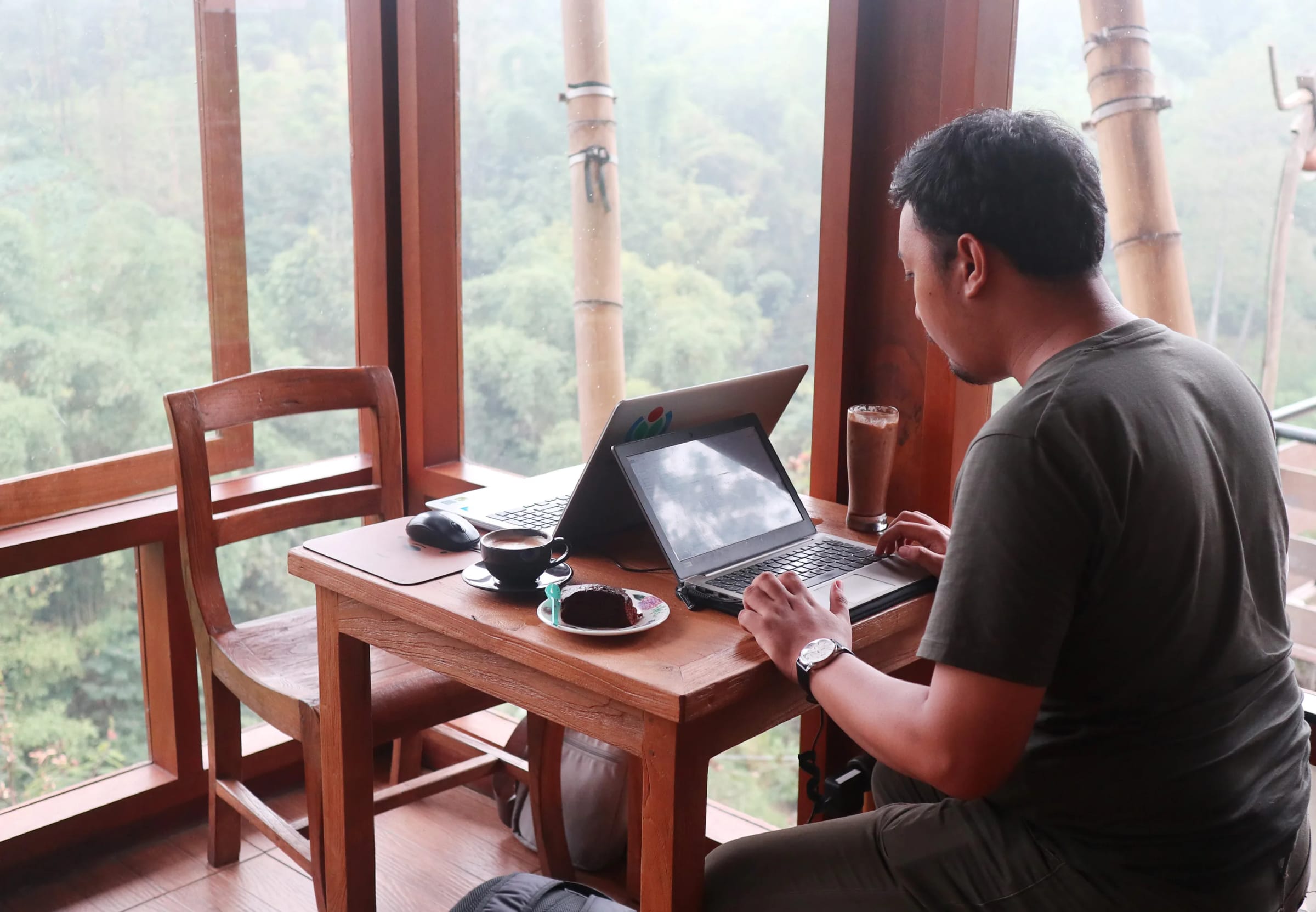 A man works on a laptop at a wooden table by the window of a café, with a forested hillside outside